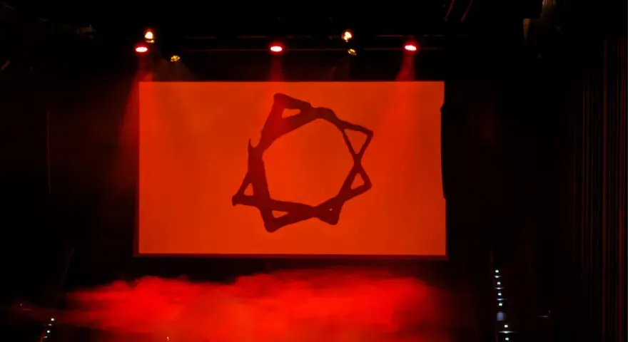 Símbolo estilizado en pantalla naranja, con humo rojo y luces de escenario. Podría ser parte de una dellafuente biografia visual.