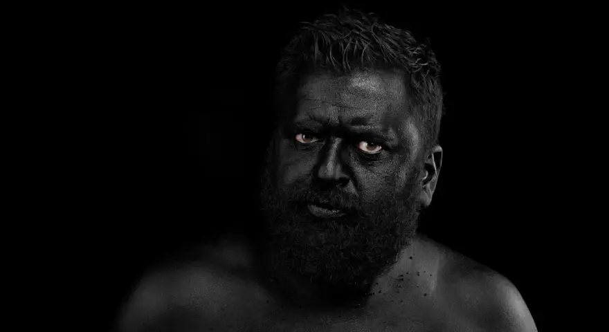 Hombre con el rostro y cuerpo cubiertos de pintura negra, ojos penetrantes. Parece un guerrero ancestral, don fluor.