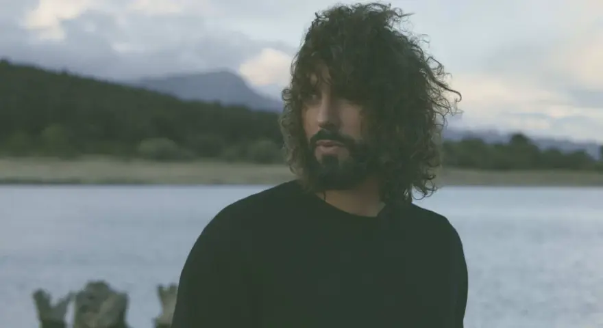 Un hombre con cabello rizado y barba mira hacia el horizonte, entre el miedo y el paraíso de un paisaje acuático y montañoso.