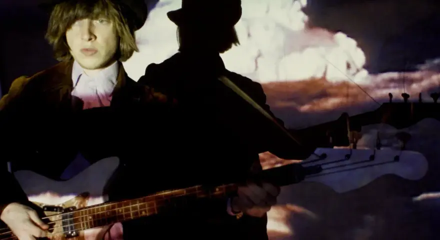 Jacco Gardner, con sombrero y chaqueta, toca el bajo frente a una proyección psicodélica.