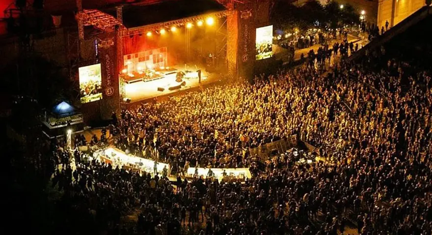 Multitudinaria asistencia a conciertos en julio. Una banda toca en un escenario iluminado ante miles de fans.