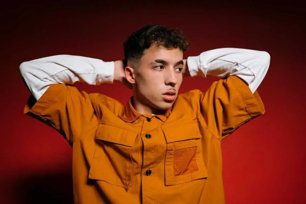 Joven con las manos detrás de la cabeza, vistiendo una chaqueta naranja y camisa blanca. Parece un pole cantante listo para actuar.