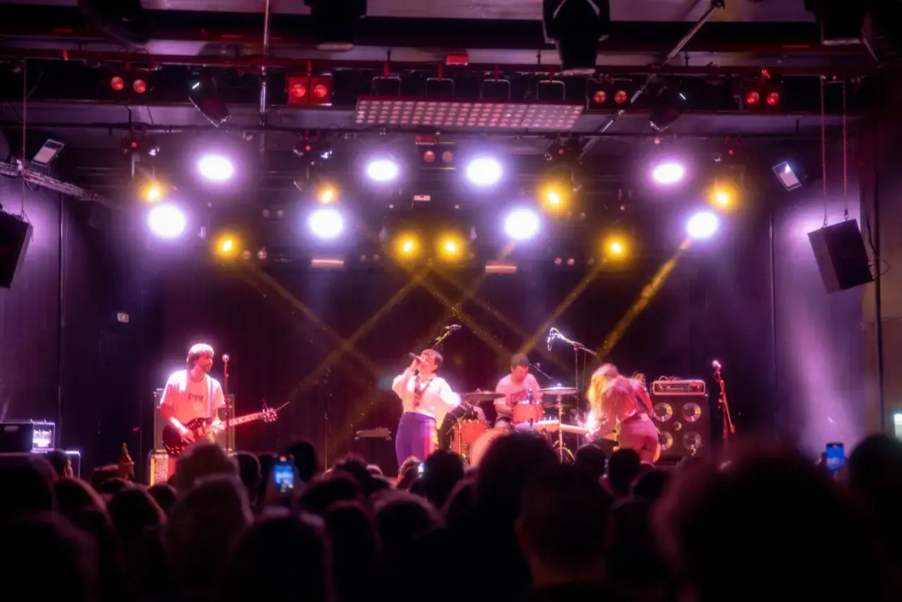 La banda Belako en concierto, con luces moradas y amarillas iluminando el escenario. La cantante principal está en el centro, con un guitarrista a su izquierda y un baterista a su derecha.