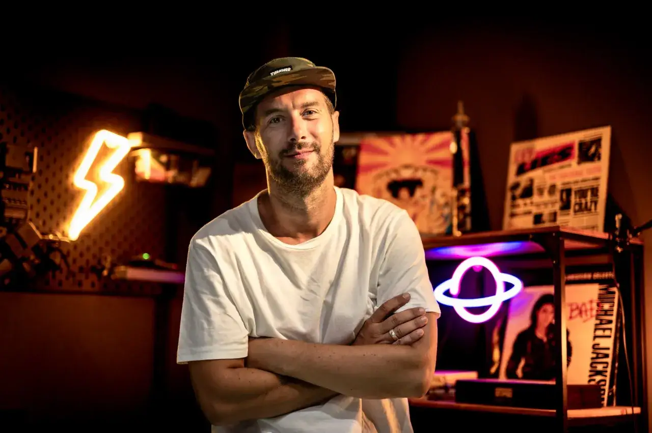 DJ Plan B, con gorra y camiseta blanca, posa con los brazos cruzados. Luces de neón y discos de vinilo de Michael Jackson de fondo.