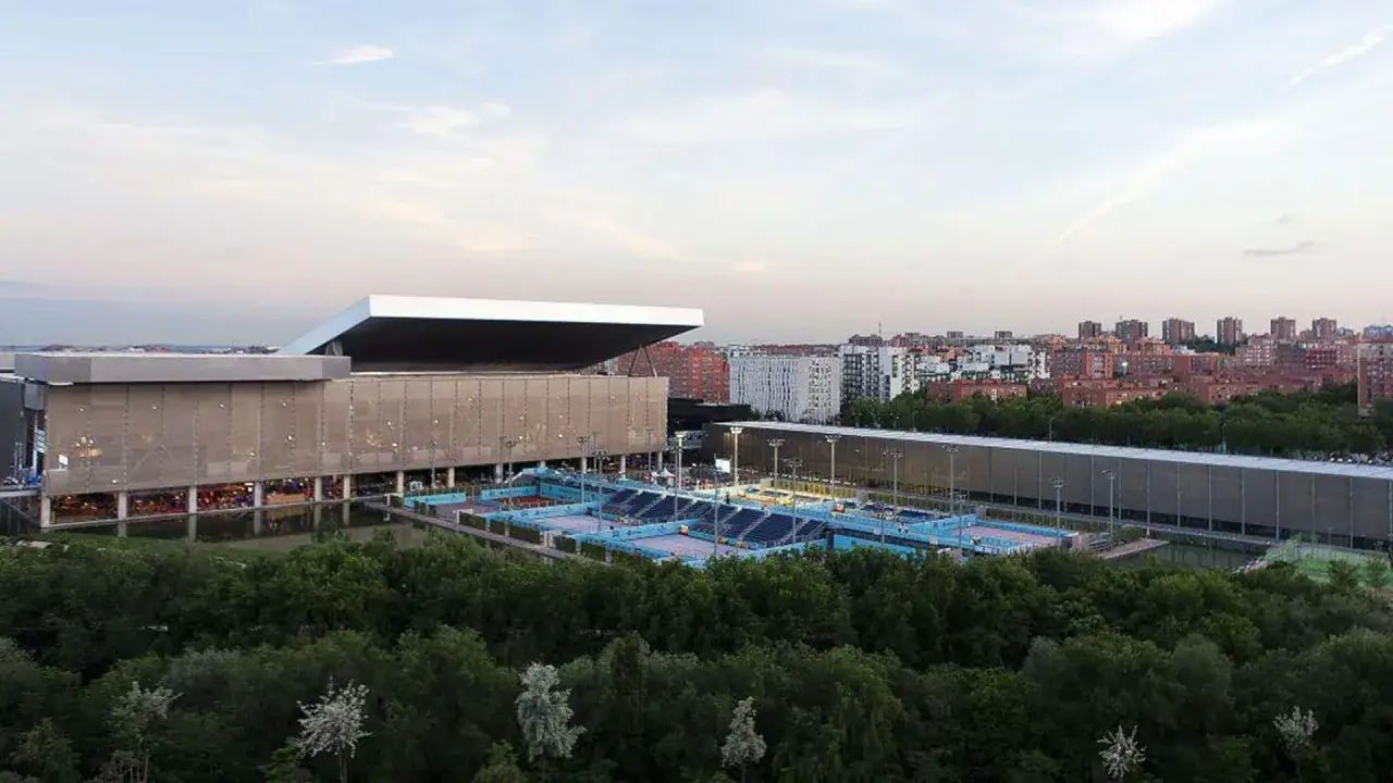 La Caja Mágica, un recinto deportivo y de ocio, se alza entre árboles y edificios. Sus pistas de tenis azules y gradas esperan a los aficionados, mientras su diseño futurista promete emocionantes conciertos.