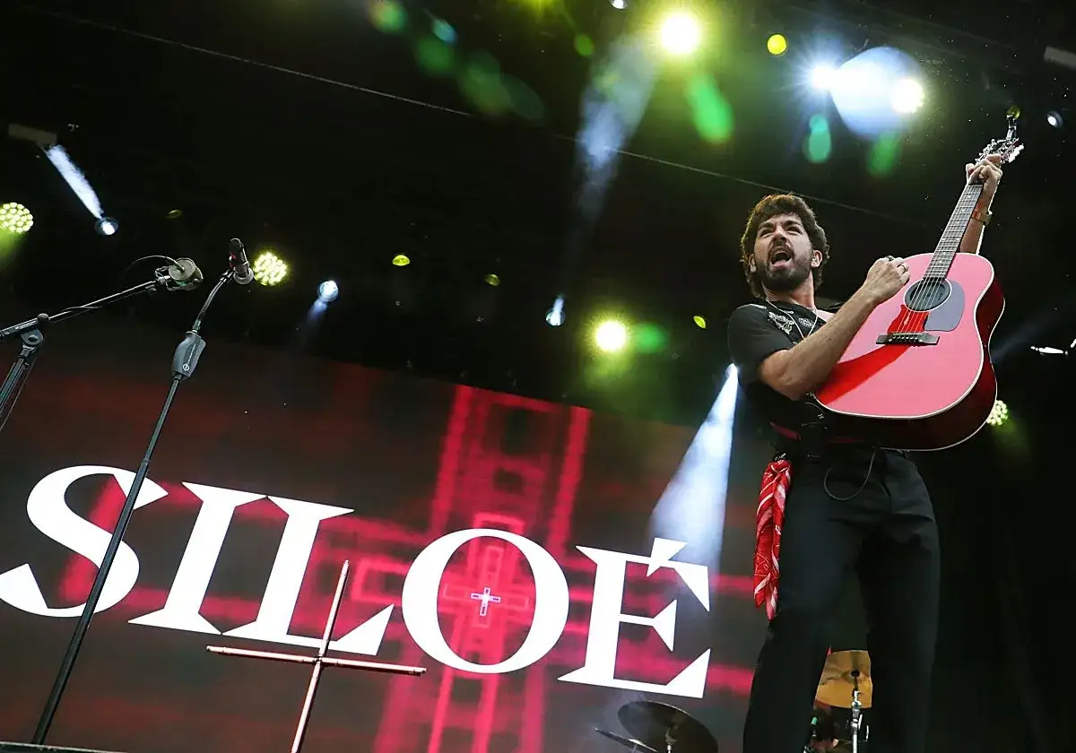 Cantante Siloe, con guitarra roja, actuando en un escenario con luces y el logo de Siloe.