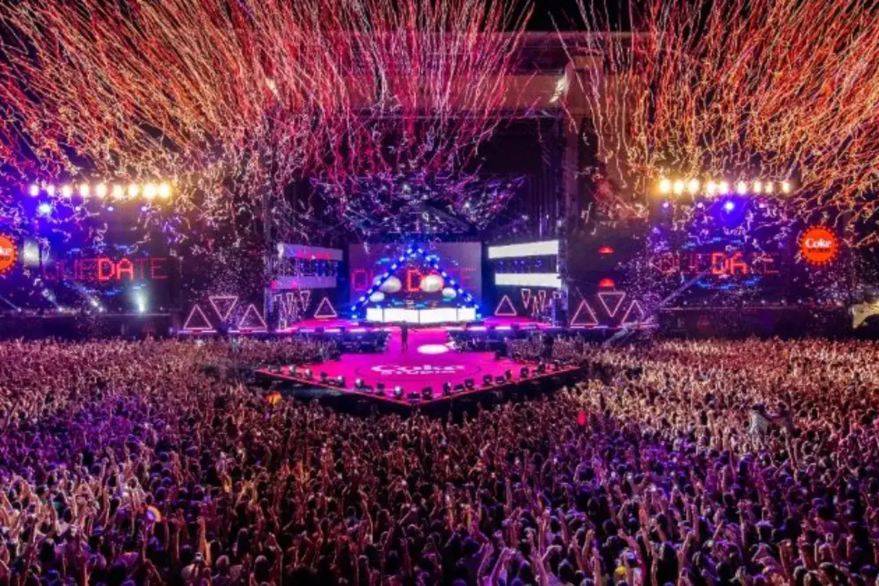 Multitud en un festival de música de Coca-Cola, con confeti rojo y morado cayendo sobre un escenario iluminado.