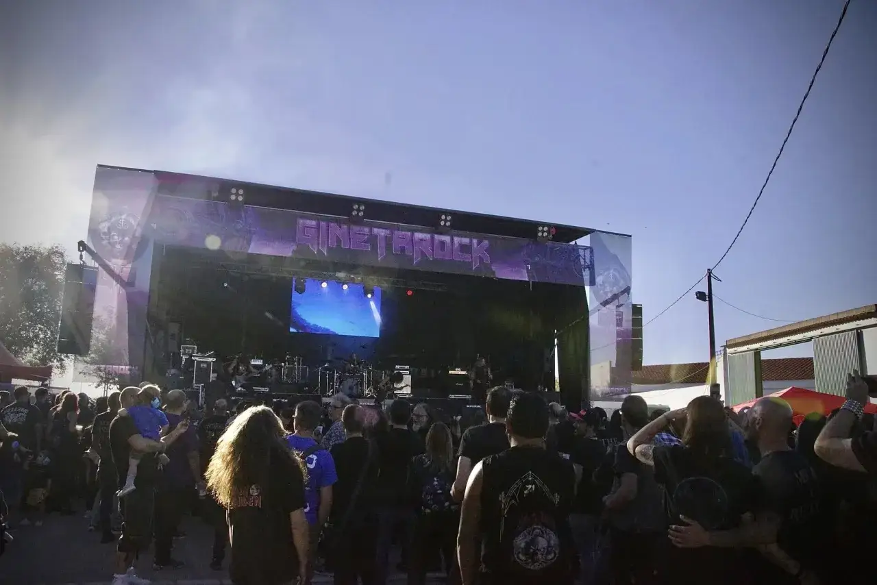 Multitud disfrutando de un concierto de gineta rock bajo un cielo azul. El escenario principal tiene el nombre del evento en letras moradas.