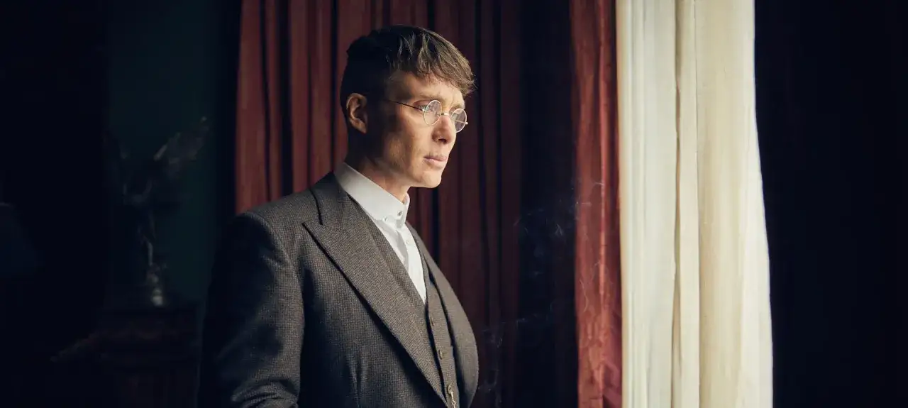 Tommy Shelby, con su icónico traje y gafas redondas, reflexiona ante la ventana. El bso Peaky Blinders resuena en el aire.