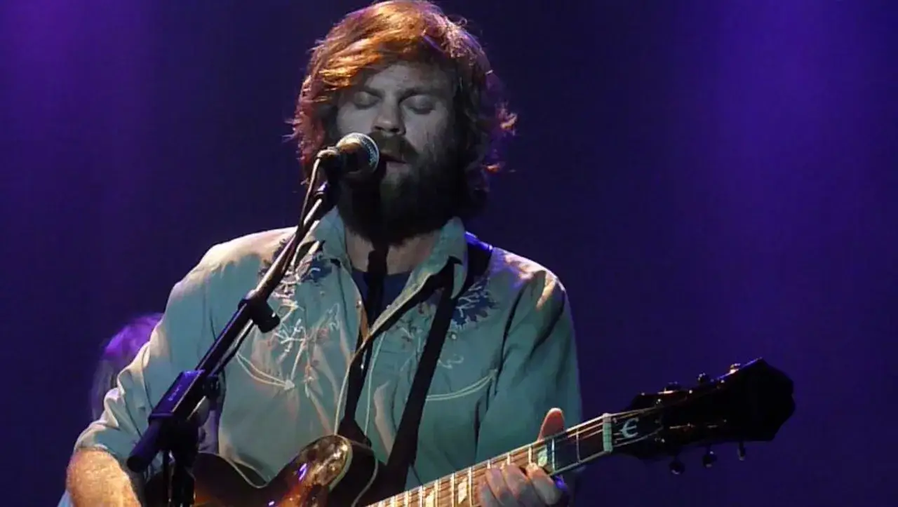 Neil Halstead, con barba y pelo largo, canta y toca la guitarra en un escenario con luces púrpuras.