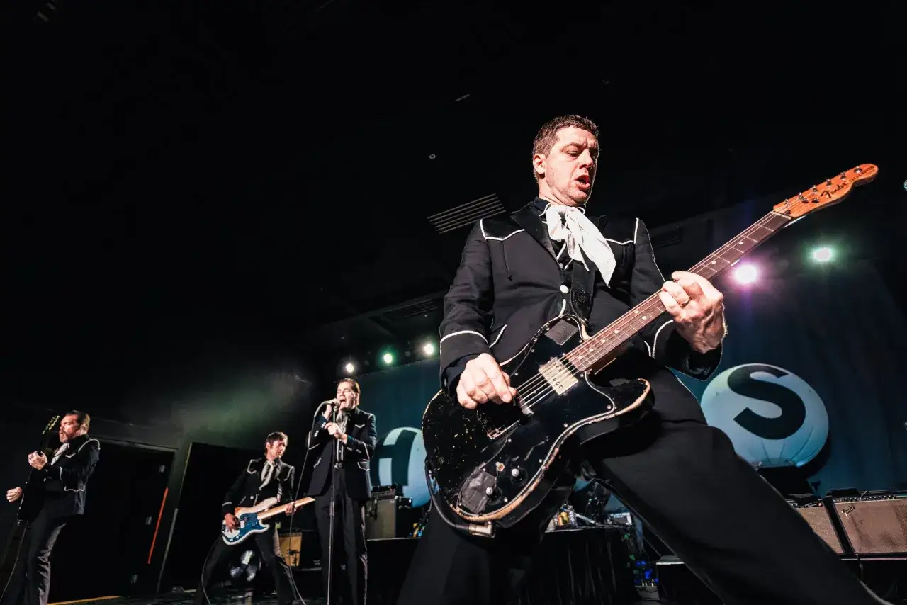 Miembros de The Hives en&eacute;rgicos en el escenario, con trajes negros y corbatas blancas, tocando guitarras el&eacute;ctricas.