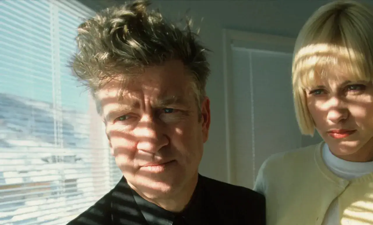 Un hombre con cabello revuelto y una mujer rubia, iluminados por sombras de persianas, parecen vivir una unrecorded night.