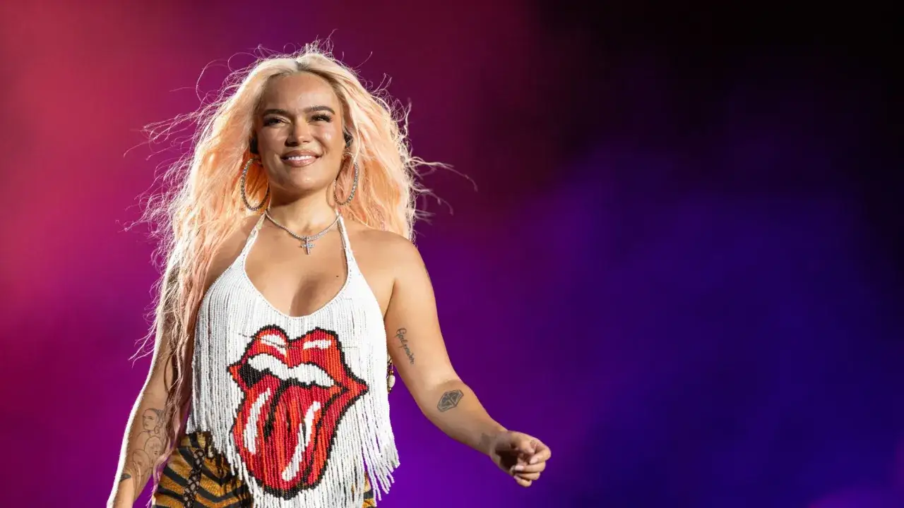 Karol G sonríe en el escenario, luciendo un top blanco con flecos y el logo de los Rolling Stones. Su cabello rubio platino ondea mientras se prepara para su gira.