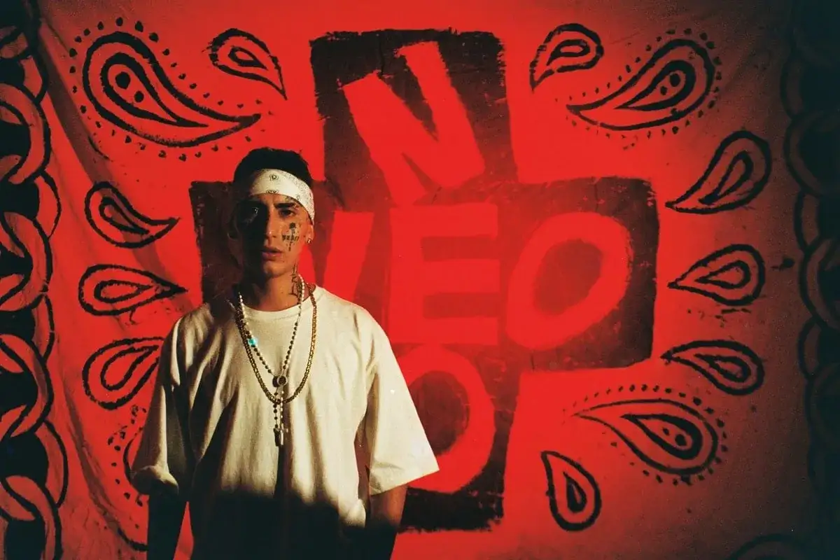 Joven con bandana y cadenas, frente a un fondo rojo con el logo