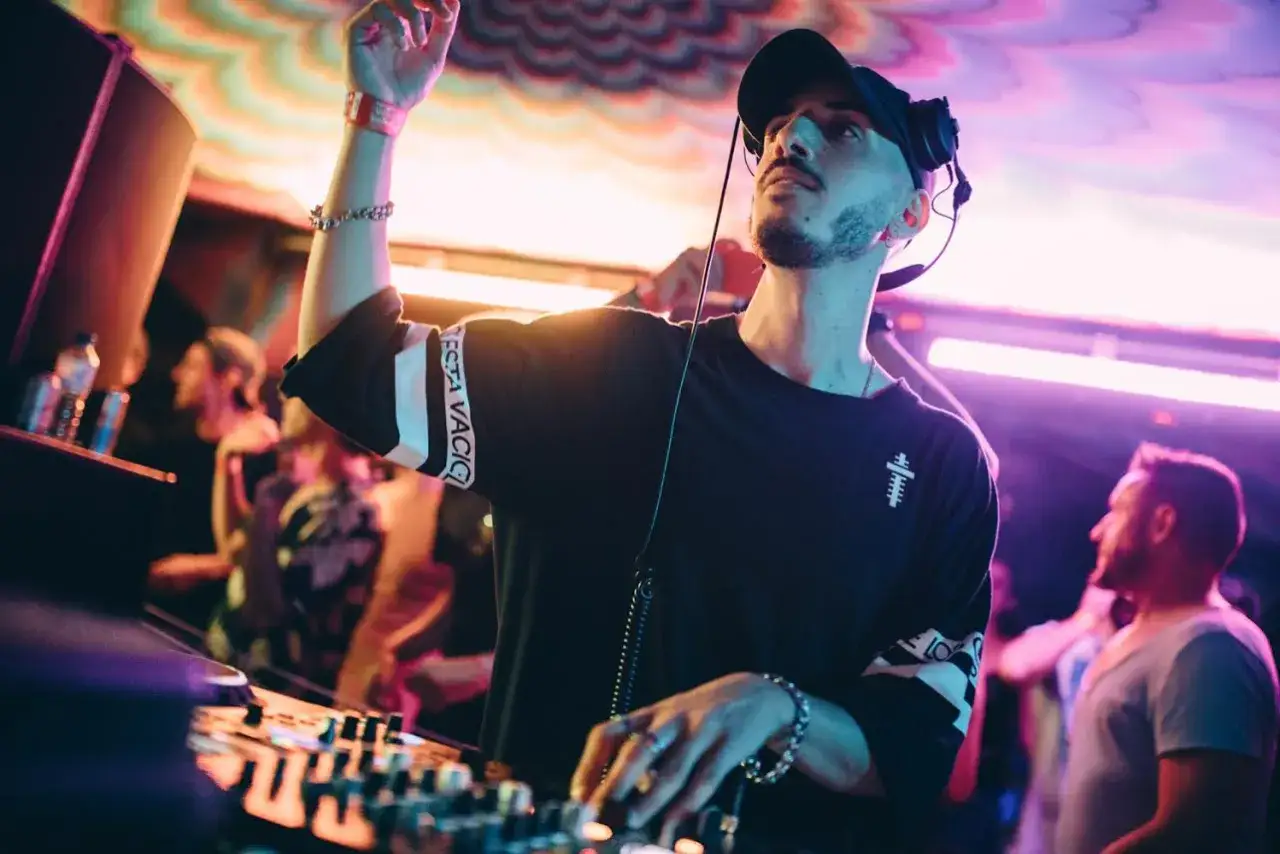 DJ wade, con auriculares y gorra, mezcla música en un club con luces de neón y público bailando.