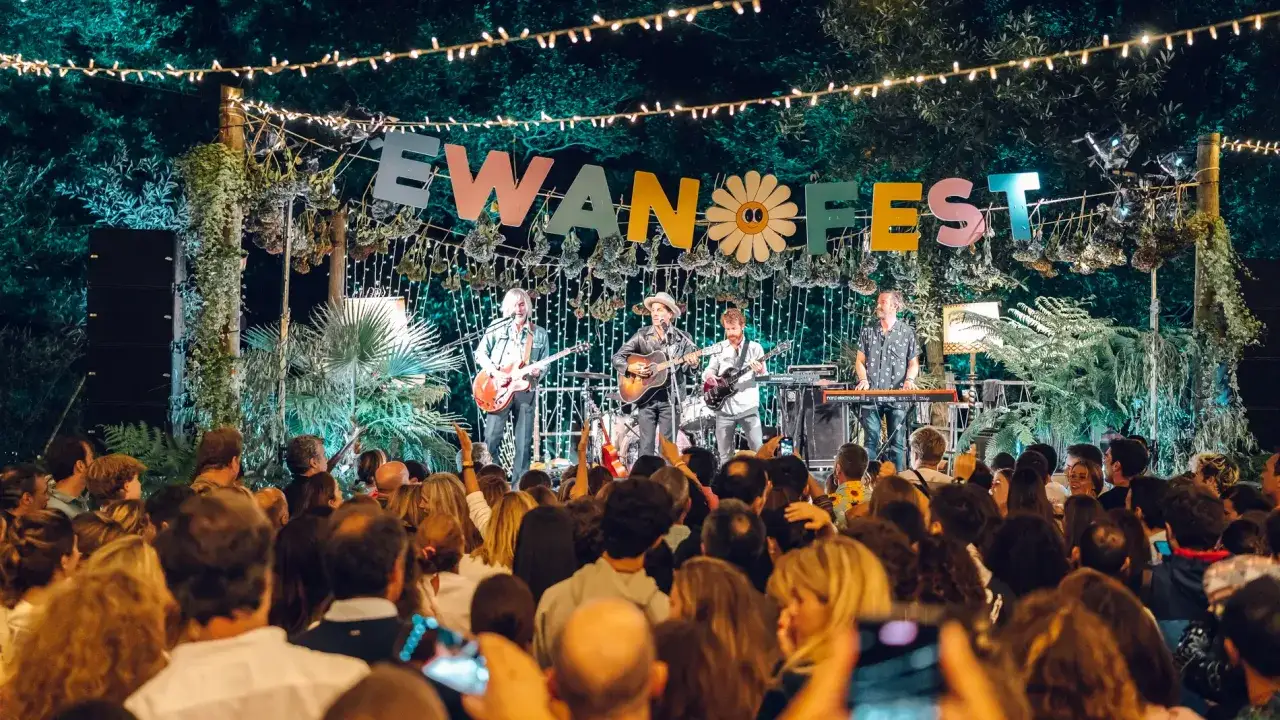 Una banda toca en el festival Ewan Salinas bajo luces y vegetación. El público disfruta del concierto.