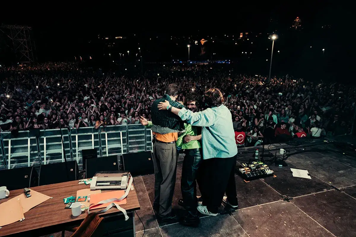 Abrazo entre músicos en un escenario ante una multitud en conciertos 1 mayo Madrid. Luces de ciudad al fondo.