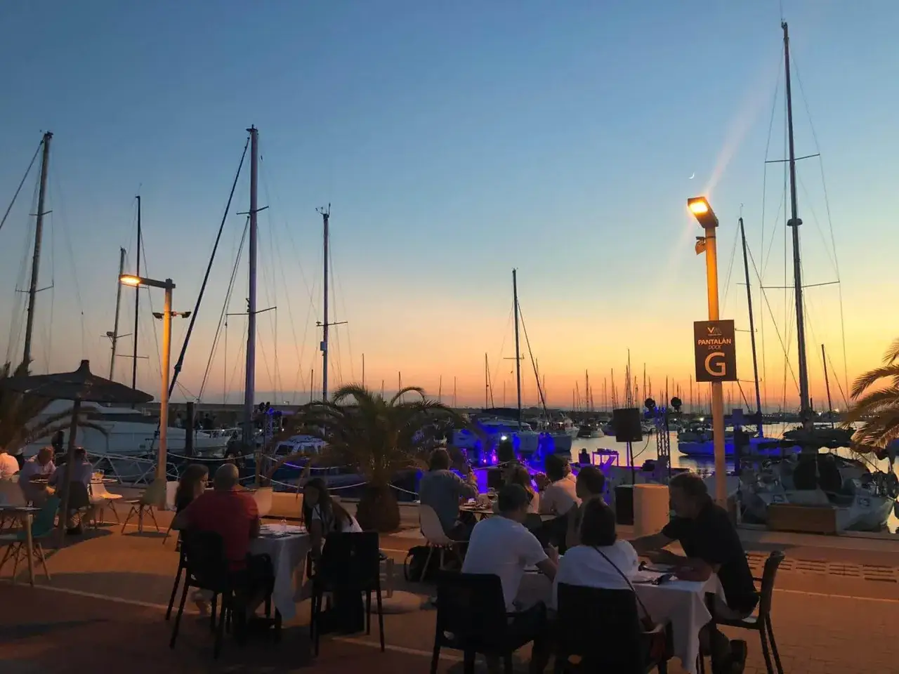 Gente disfrutando de una cena al aire libre en un puerto deportivo al atardecer, con veleros y luces de colores. ¡Me encanta rockear en Valencia!