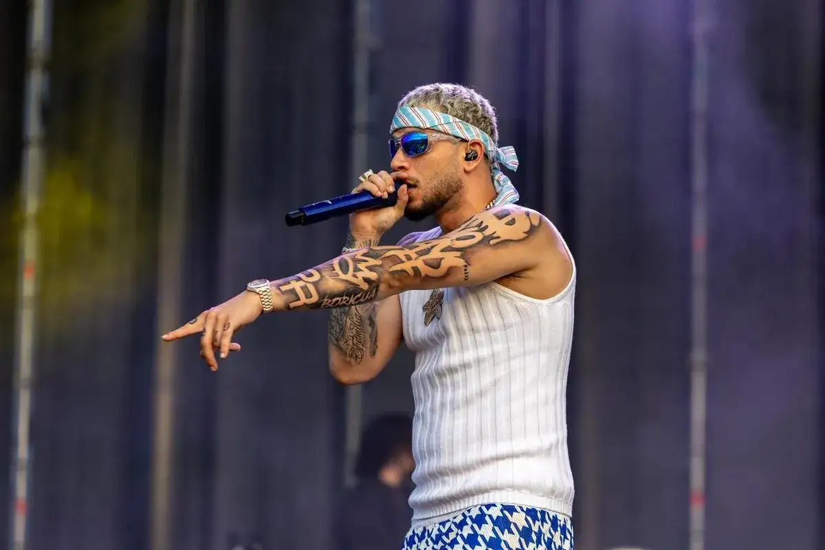 Artista con bandana y gafas de sol canta en un concierto en Málaga. Sus brazos están cubiertos de tatuajes, incluyendo