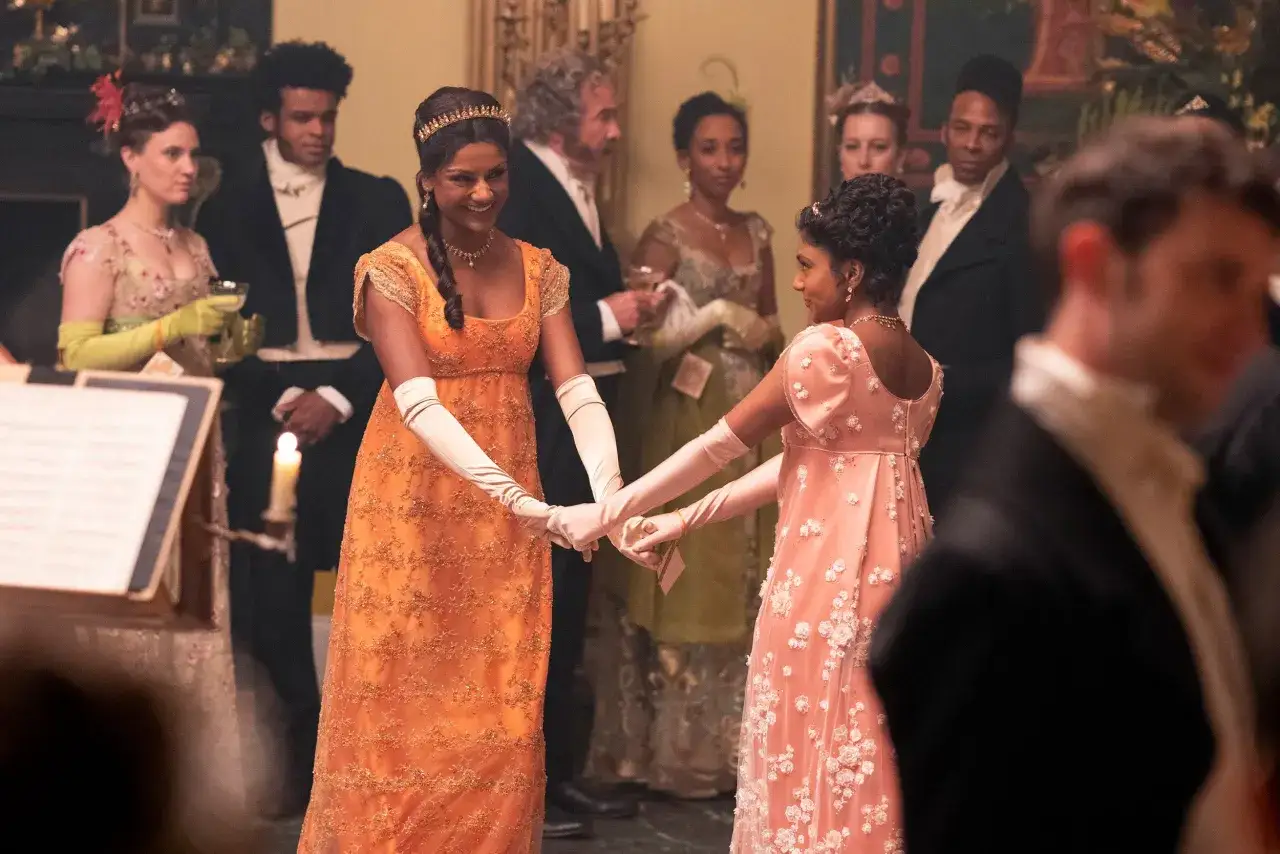 Dos damas con vestidos de época, una naranja y otra rosa, se toman de las manos en un salón de baile. La escena evoca la banda sonora de los Bridgerton.