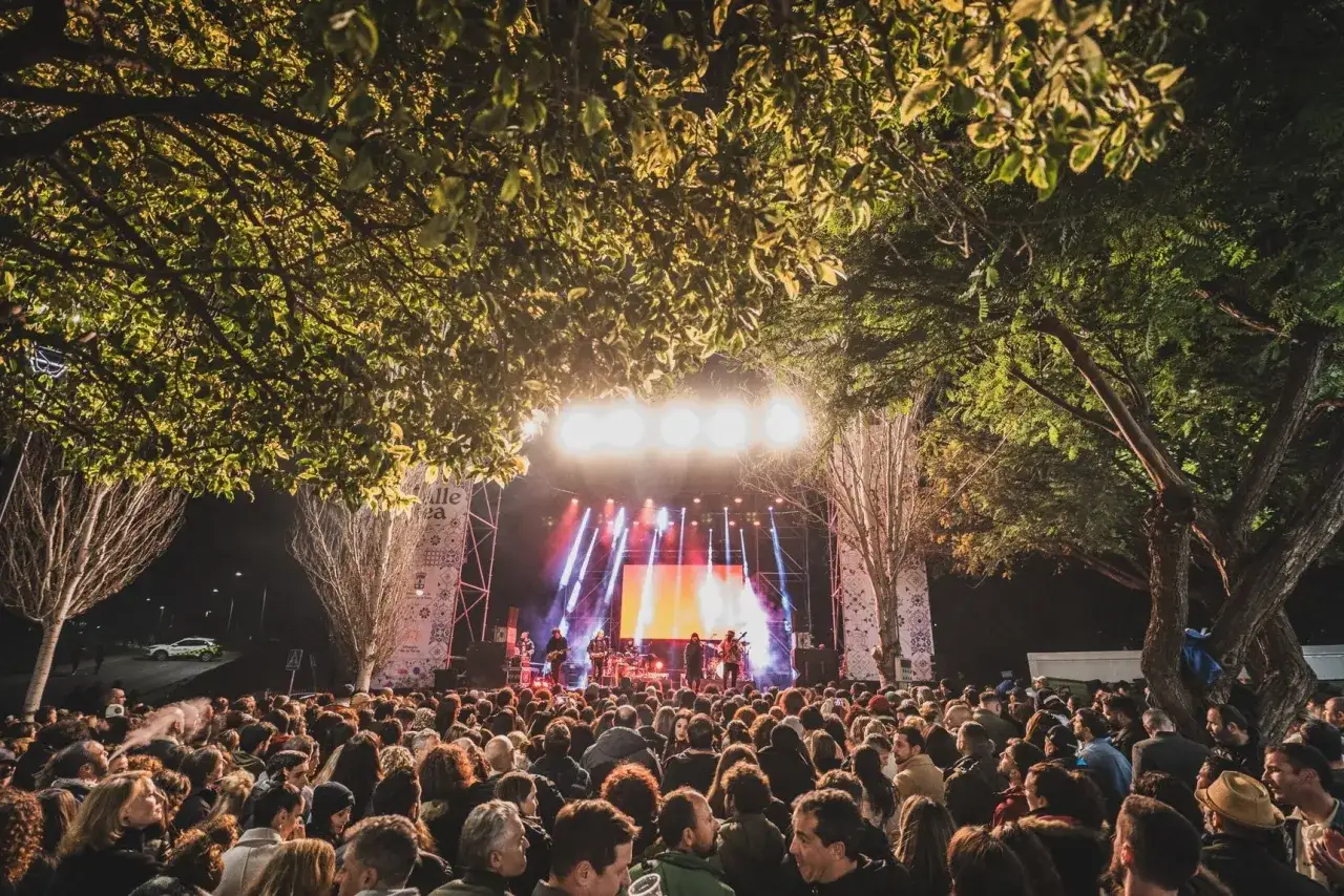 Multitud disfrutando de un concierto en el Sophie Festival bajo árboles frondosos. Luces brillantes iluminan el escenario.