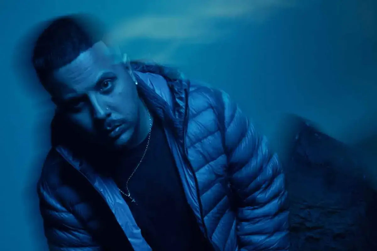 Joven con chaqueta azul, collar con cruz y piercing. El cruz cafuné significado se refleja en su mirada introspectiva.