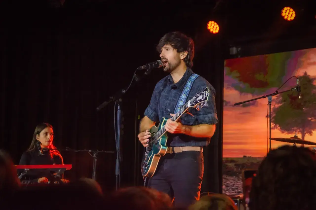 Músico canta y toca guitarra en un escenario con una tecladista al fondo. La pantalla proyecta un atardecer, como si fuera el mar donde nada el pescao.