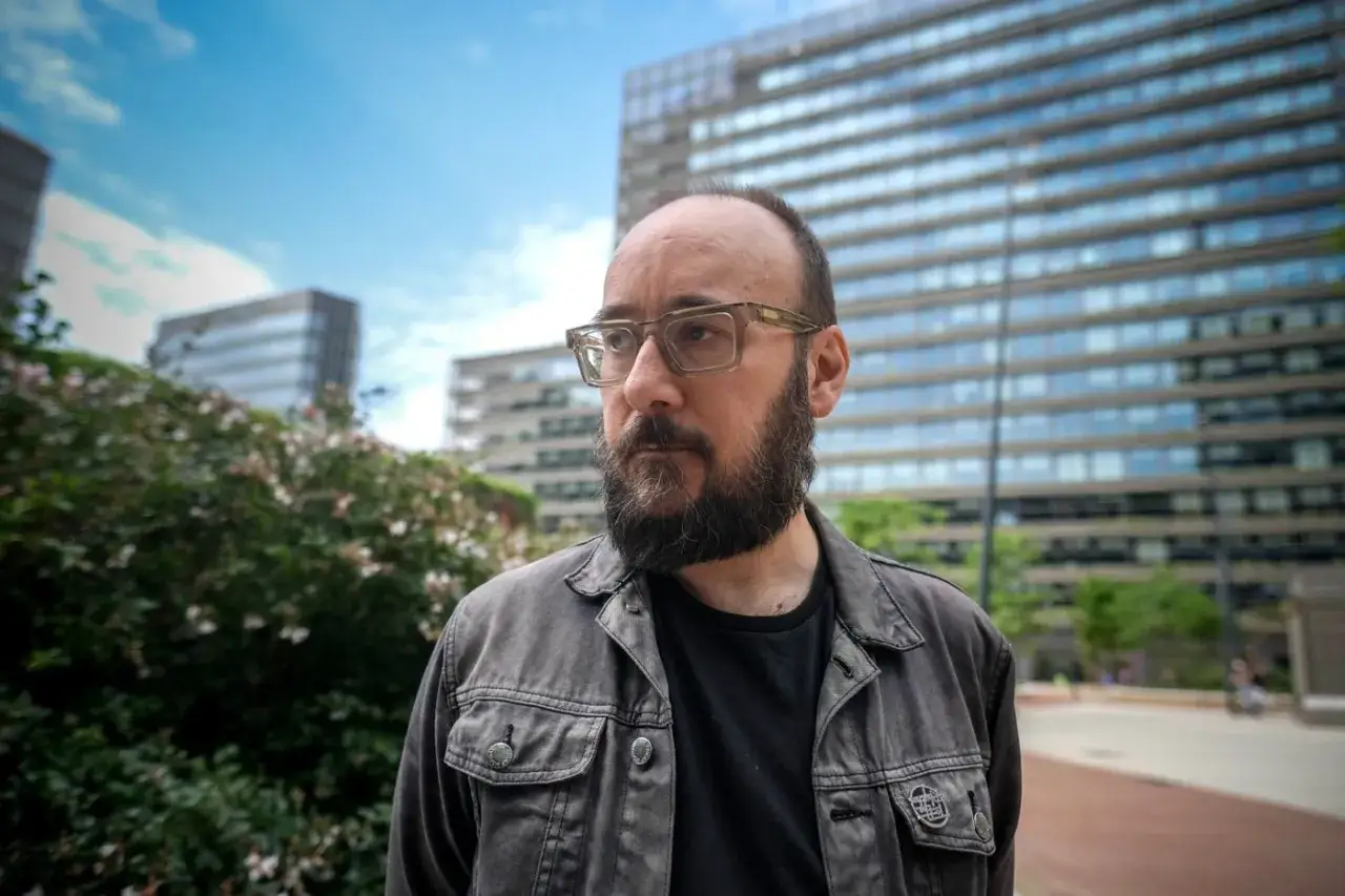 Dj Coco, con gafas y barba, posa frente a edificios modernos y vegetación exuberante.