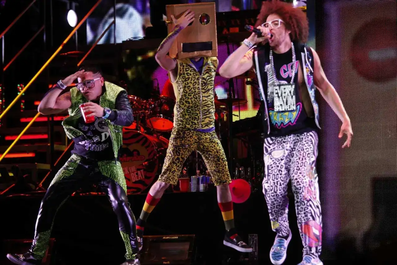 Los miembros de LMFAO en el escenario, uno con un disfraz de caja en la cabeza, otro cantando y el tercero con un vaso rojo.
