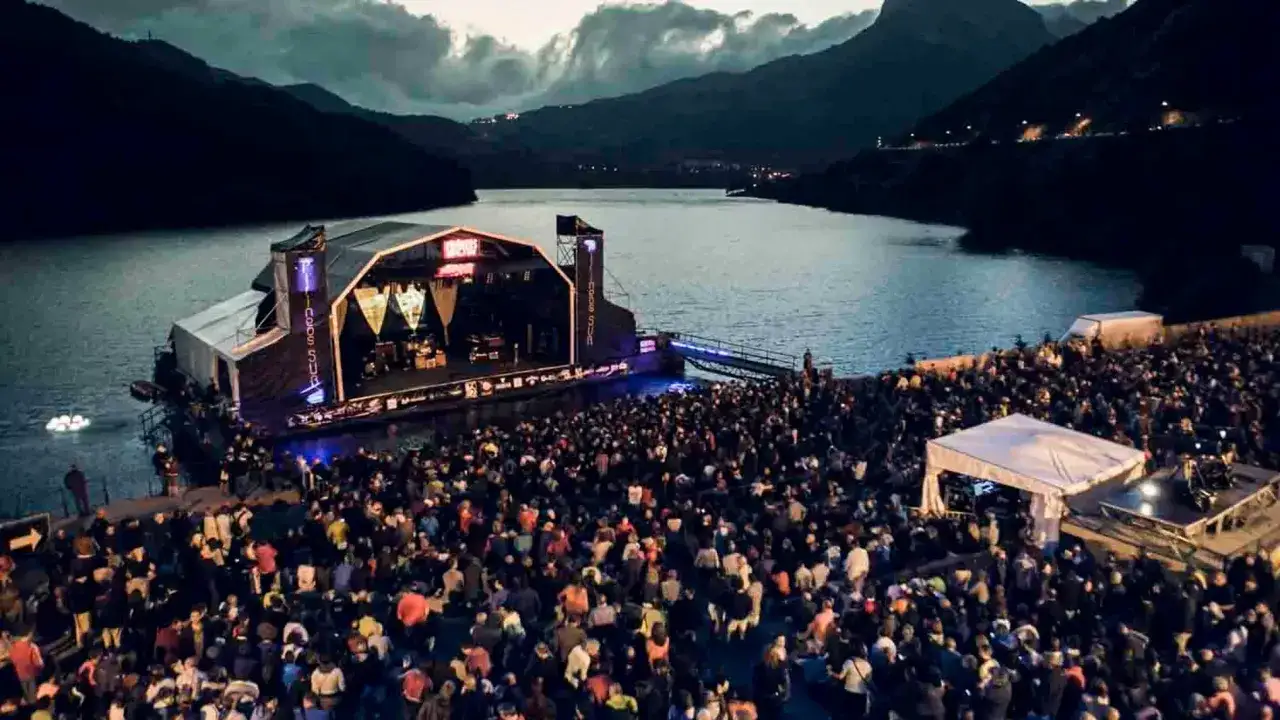 Multitud disfrutando de un concierto en Lollapalooza España, con un escenario flotante sobre un lago y montañas al fondo.