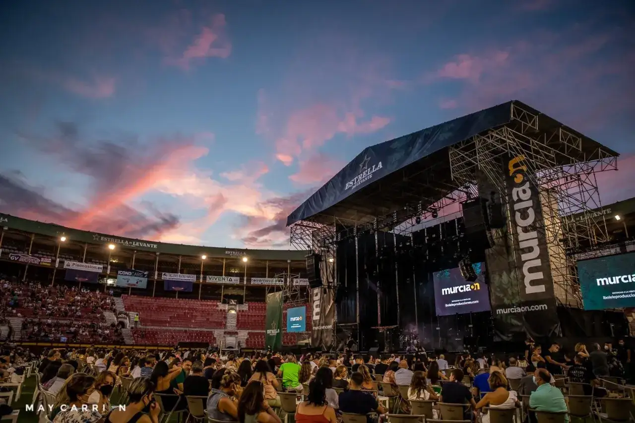 Un concierto en Murcia bajo un cielo rosado. El público espera la actuación en un recinto con el logo de murciaon.