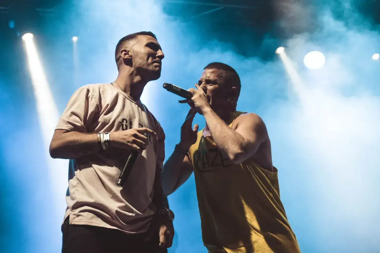 Dos cantantes en el escenario, uno con camiseta rosa y otro con camiseta amarilla con la palabra