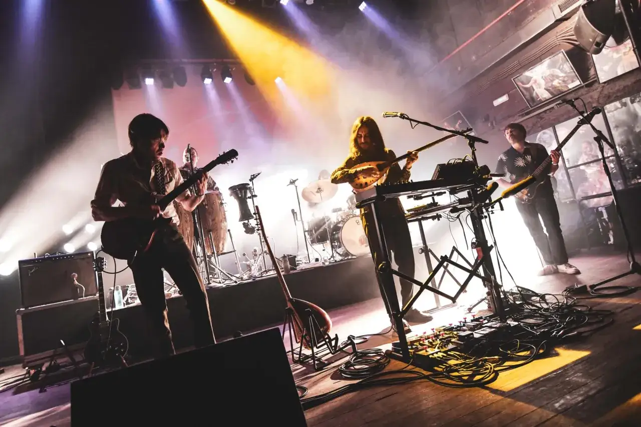 Banda tocando en vivo, con guitarras, teclados y percusión. La iluminación crea una atmósfera de concierto, como si los track dogs estuvieran persiguiendo el ritmo.