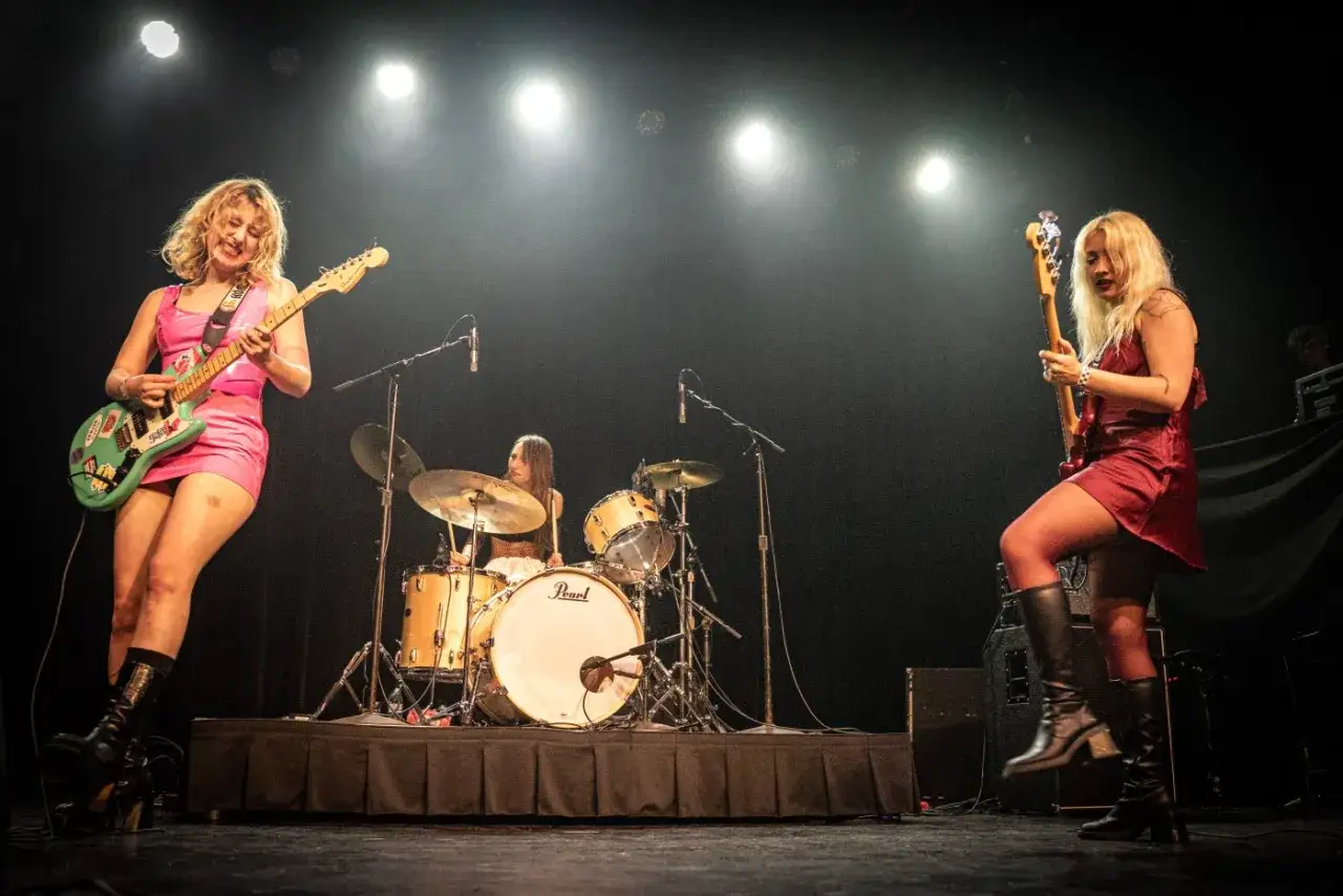 Tres mujeres enérgicas en el escenario, una tocando la guitarra eléctrica verde, otra la batería y la tercera el bajo. Puro rock en femenino.