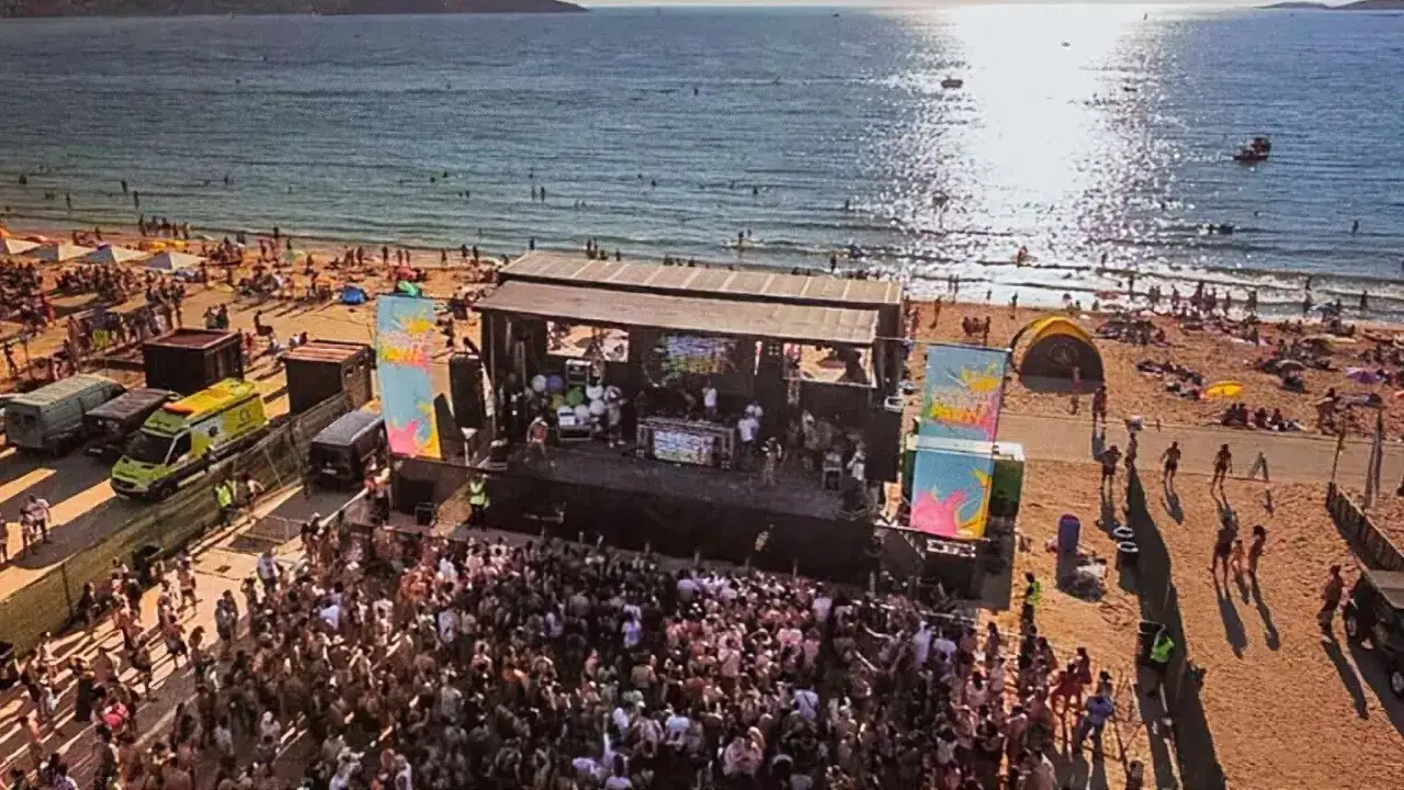 Multitud bailando en un festival de música en la playa al atardecer. El sol brilla sobre el mar, con gente disfrutando del agua y la arena.