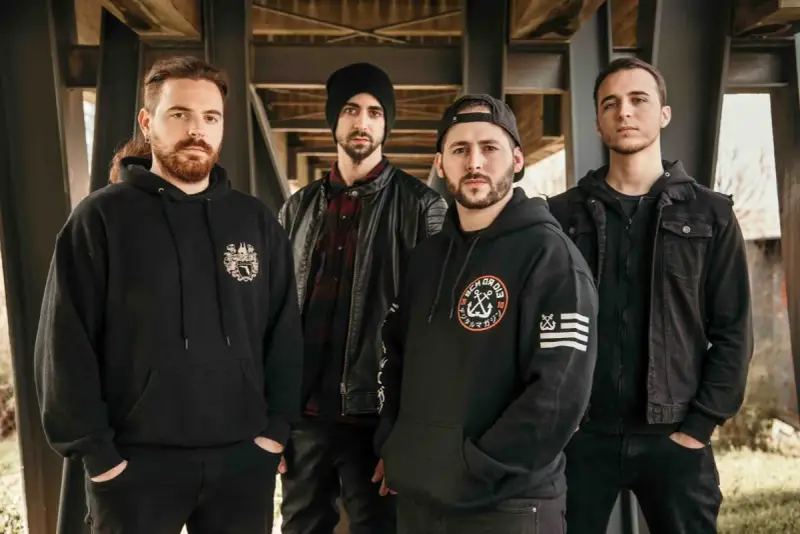 Cuatro hombres con sudaderas y chaquetas negras posan bajo una estructura metálica. Sus miradas intensas parecen **blaze out** con determinación.
