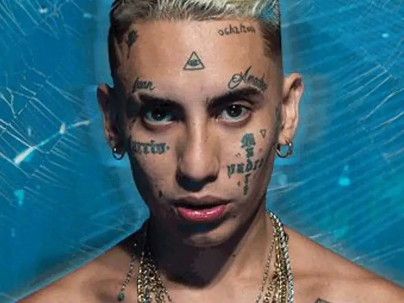 Joven con tatuajes en la cara, incluyendo un ojo dentro de un triángulo, y cadenas de oro. Su estilo neo pistea madrid es inconfundible.
